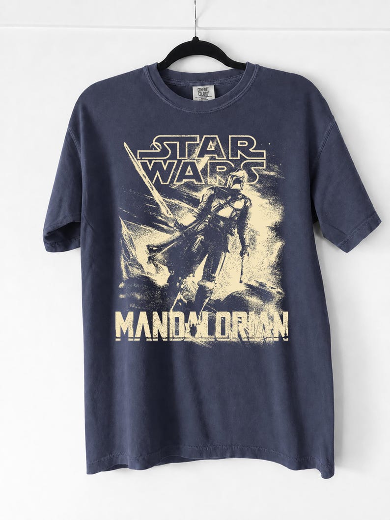 May include: Navy blue t-shirt featuring a distressed graphic of The Mandalorian from Star Wars. The design includes the Star Wars logo and the word "Mandalorian" in a vintage style. The shirt is hanging on a black hanger.