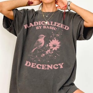 Könnte beinhalten: Ein anthrazitfarbenes Kurzarm-T-Shirt mit einer verblassten rosa Grafik eines Vogels auf einem blühenden Zweig. Der Text RADICALIZED BY BASIC DECENCY ist in einer abgenutzten rosa Schrift über und unter der Vogelillustration gedruckt.