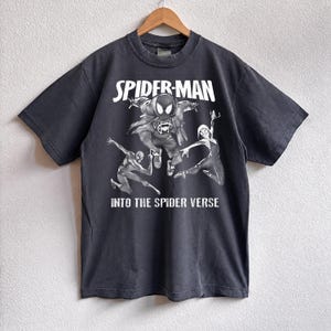 May include: Dark gray t-shirt featuring a graphic from the movie "Spider-Man: Into the Spider-Verse." The design includes the words "Spider-Man" and "Into the Spider Verse" with illustrations of Spider-Man characters.