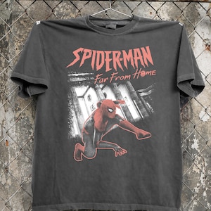 K&ouml;nnte beinhalten: Dunkelgraues T-Shirt mit rotem Schriftzug "SPIDER-MAN Far From Home". Die Grafik zeigt Spider-Man in einem rot-schwarzen Anzug, der vor einem Geb&auml;ude kniet. Das T-Shirt h&auml;ngt an einem Metallb&uuml;gel.