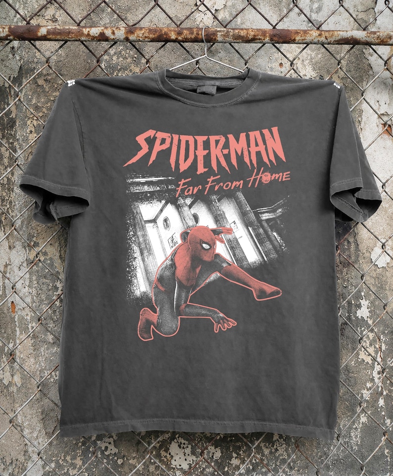 K&ouml;nnte beinhalten: Dunkelgraues T-Shirt mit rotem Schriftzug "SPIDER-MAN Far From Home". Die Grafik zeigt Spider-Man in einem rot-schwarzen Anzug, der vor einem Geb&auml;ude kniet. Das T-Shirt h&auml;ngt an einem Metallb&uuml;gel.