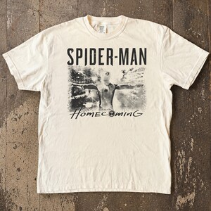 May include: Cream-colored t-shirt featuring the text "SPIDER-MAN" above a black and white image of Spider-Man with the word "Homecoming" below. The shirt has short sleeves and a crew neck.