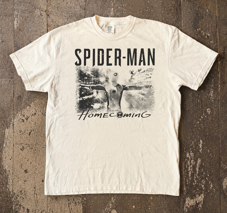 May include: Cream-colored t-shirt featuring the text "SPIDER-MAN" above a black and white image of Spider-Man with the word "Homecoming" below. The shirt has short sleeves and a crew neck.