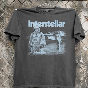 Peut inclure: T-shirt gris foncé avec le mot "Interstellar" en bleu clair au-dessus d'un graphisme de deux astronautes. Un astronaute est debout dans l'eau, et l'autre marche vers un vaisseau spatial. Le t-shirt est suspendu à une clôture métallique.