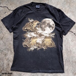 May include: A black short-sleeve t-shirt featuring a graphic print of three brown toads positioned around a large, detailed white full moon. The design includes cloudy, textured elements surrounding the toads and the moon on the front of the shirt.
