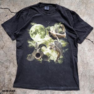 May include: A black short-sleeve t-shirt featuring a graphic print of three fish swimming against a backdrop of a large, glowing full moon and green nebulous clouds. The fish are depicted in various positions with detailed scales and open mouths.