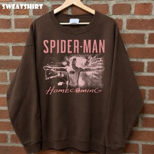 May include: Brown sweatshirt featuring the words "SPIDER-MAN" and "Homecoming" in pink lettering. The graphic depicts Spider-Man with outstretched arms. The word "SWEATSHIRT" is printed at the top of the image.