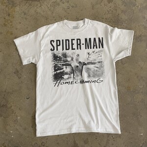 May include: White t-shirt featuring a graphic of Spider-Man from the movie "Homecoming." The design is in black and white, with the words "SPIDER-MAN" above the image and "Homecoming" below. The shirt has a crew neck and short sleeves.