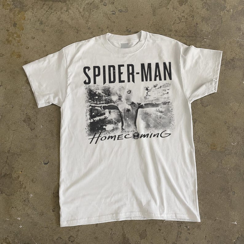 May include: White t-shirt featuring a graphic of Spider-Man from the movie "Homecoming." The design is in black and white, with the words "SPIDER-MAN" above the image and "Homecoming" below. The shirt has a crew neck and short sleeves.