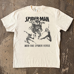 May include: Cream-colored t-shirt featuring a black and white graphic of Spider-Man and other Spider-Verse characters. The text "SPIDER-MAN" is above the graphic, and "INTO THE SPIDER VERSE" is below.