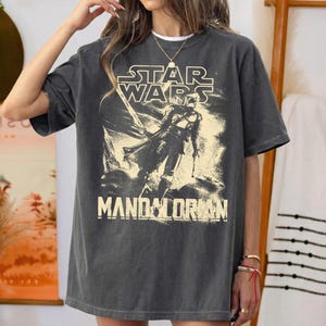 May include: Dark gray t-shirt featuring a Star Wars graphic with the text "STAR WARS" above an illustration of The Mandalorian. The text "MANDALORIAN" is printed below the illustration. The shirt has a vintage, worn look.