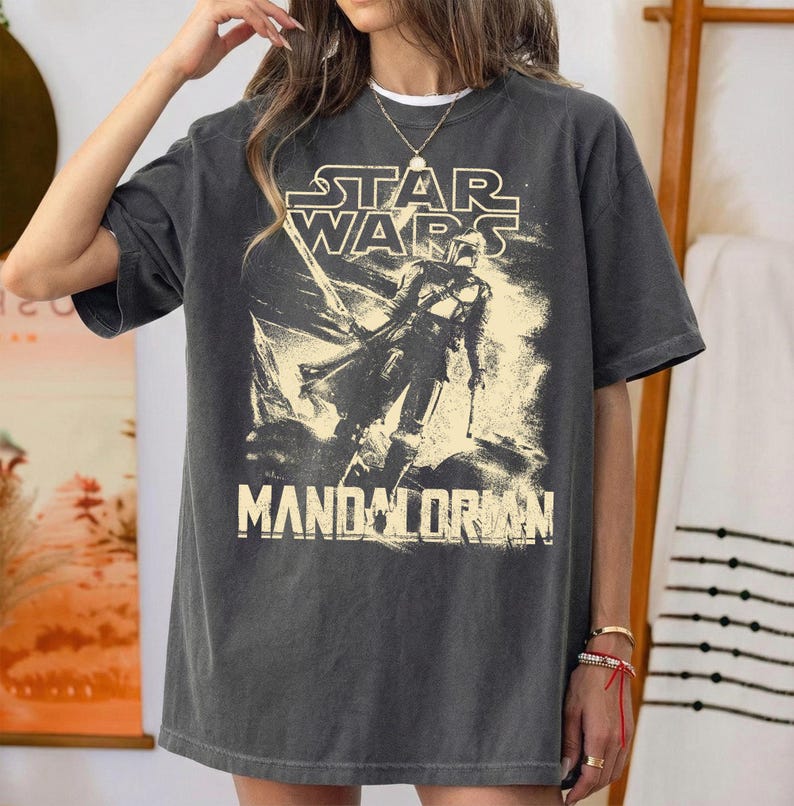 May include: Dark gray t-shirt featuring a Star Wars graphic with the text "STAR WARS" above an illustration of The Mandalorian. The text "MANDALORIAN" is printed below the illustration. The shirt has a vintage, worn look.