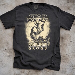 May include: Dark gray t-shirt featuring a graphic of The Mandalorian holding a blaster, with Grogu in the background. The Star Wars logo is at the top, and "The Mandalorian and Grogu" is printed below the image.