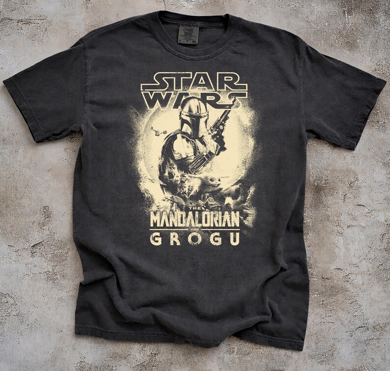 May include: Dark gray t-shirt featuring a graphic of The Mandalorian holding a blaster, with Grogu in the background. The Star Wars logo is at the top, and "The Mandalorian and Grogu" is printed below the image.