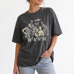 Może przedstawiać: Ciemnoszary, kwasowo prany t-shirt z krótkim rękawem, ozdobiony grafiką w stylu vintage przedstawiającą obsadę serialu M*A*S*H. Wzór zawiera tytuł serialu białymi literami na dole oraz znak drogowy w tle za postaciami.