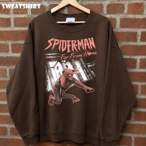 K&ouml;nnte beinhalten: Braunes Sweatshirt mit einer Grafik von Spider-Man aus dem Film "Far From Home". Das Design zeigt Spider-Man in einer hockenden Pose mit einem Stadtlandschaftshintergrund. Das Wort "SWEATSHIRT" steht oben.