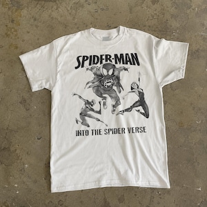 May include: White t-shirt featuring a graphic of Spider-Man characters from the movie "Into the Spider-Verse." The design includes the words "SPIDER-MAN" above the characters and "INTO THE SPIDER VERSE" below. The shirt has a crew neck and short sleeves.