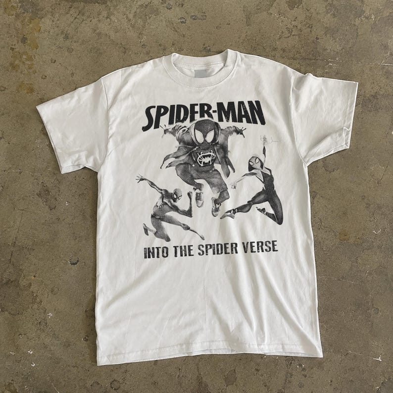 May include: White t-shirt featuring a graphic of Spider-Man characters from the movie "Into the Spider-Verse." The design includes the words "SPIDER-MAN" above the characters and "INTO THE SPIDER VERSE" below. The shirt has a crew neck and short sleeves.