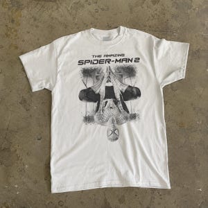 May include: White t-shirt featuring a black and white graphic of Spider-Man hanging upside down. The text "THE AMAZING SPIDER-MAN 2" is printed above the graphic. The shirt has short sleeves and a crew neck.