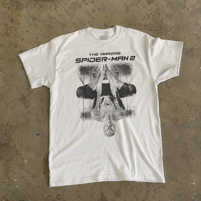 May include: White t-shirt featuring a black and white graphic of Spider-Man hanging upside down. The text "THE AMAZING SPIDER-MAN 2" is printed above the graphic. The shirt has short sleeves and a crew neck.
