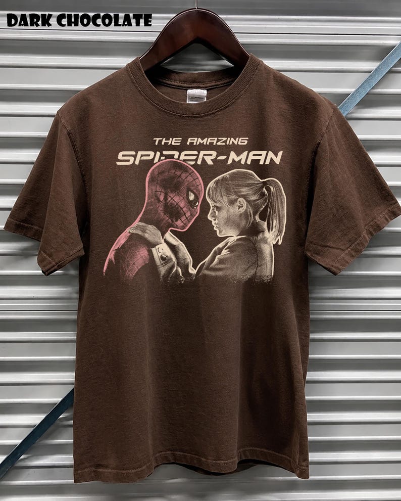 K&ouml;nnte beinhalten: Dunkel schokoladenbraunes T-Shirt mit einem Grafikmotiv aus "The Amazing Spider-Man". Das Design zeigt Spider-Man und eine Frau, die sich gegen&uuml;berstehen. Der Text "THE AMAZING SPIDER-MAN" ist &uuml;ber der Grafik aufgedruckt. Das T-Shirt h&auml;ngt an einem Holzkleiderb&uuml;gel.