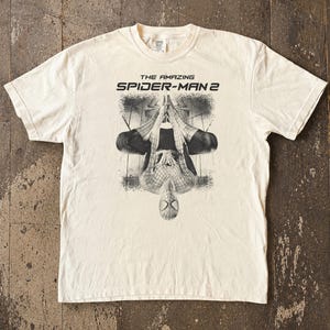 May include: Cream-colored t-shirt featuring a black and white graphic of Spider-Man from "The Amazing Spider-Man 2." The design shows Spider-Man upside down. The text "THE AMAZING SPIDER-MAN 2" is printed above the graphic.