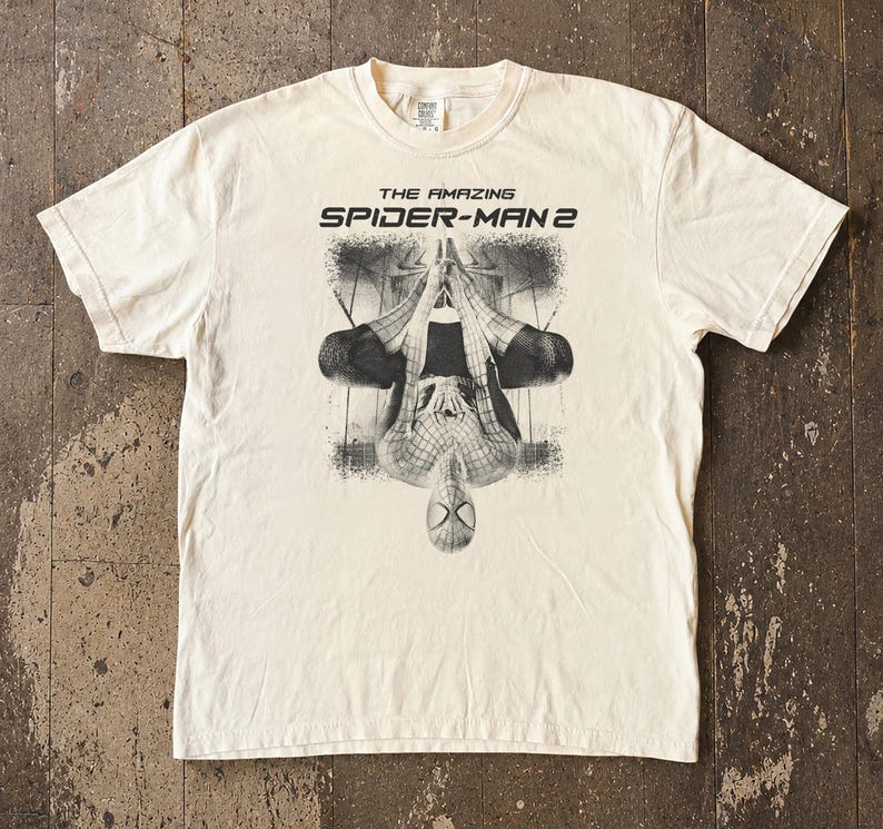 May include: Cream-colored t-shirt featuring a black and white graphic of Spider-Man from "The Amazing Spider-Man 2." The design shows Spider-Man upside down. The text "THE AMAZING SPIDER-MAN 2" is printed above the graphic.