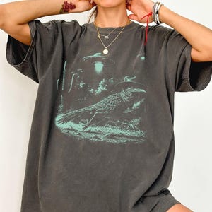 Peut inclure: Un t-shirt gris anthracite à manches courtes avec une impression graphique vert clair représentant un oiseau portant un chapeau de cowboy dans un paysage désertique avec un cactus et une soucoupe volante en arrière-plan.