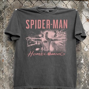 May include: Dark gray t-shirt featuring the text "SPIDER-MAN" in pink above an image of Spider-Man with outstretched arms. The word "Homecoming" is printed below the image in pink.