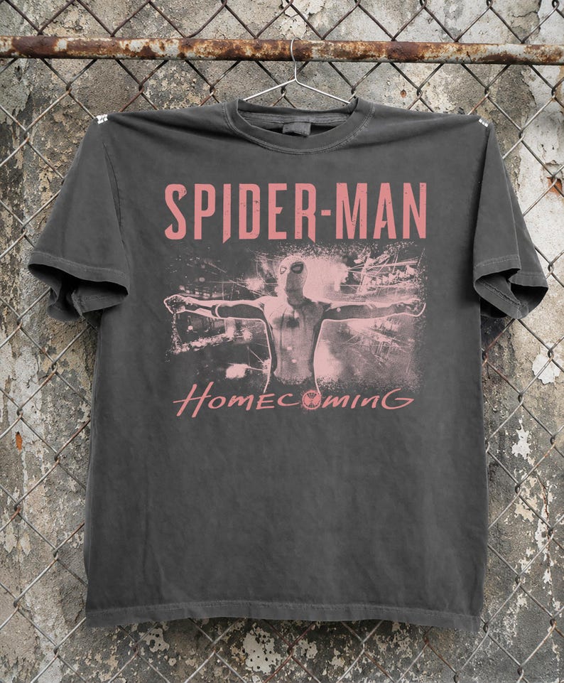 May include: Dark gray t-shirt featuring the text "SPIDER-MAN" in pink above an image of Spider-Man with outstretched arms. The word "Homecoming" is printed below the image in pink.
