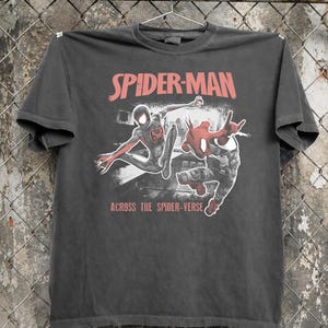 Puede incluir: Camiseta gris oscuro con la inscripci&oacute;n roja "SPIDER-MAN" y un gr&aacute;fico de dos personajes de Spider-Man. La frase "ACROSS THE SPIDER-VERSE" est&aacute; impresa debajo.