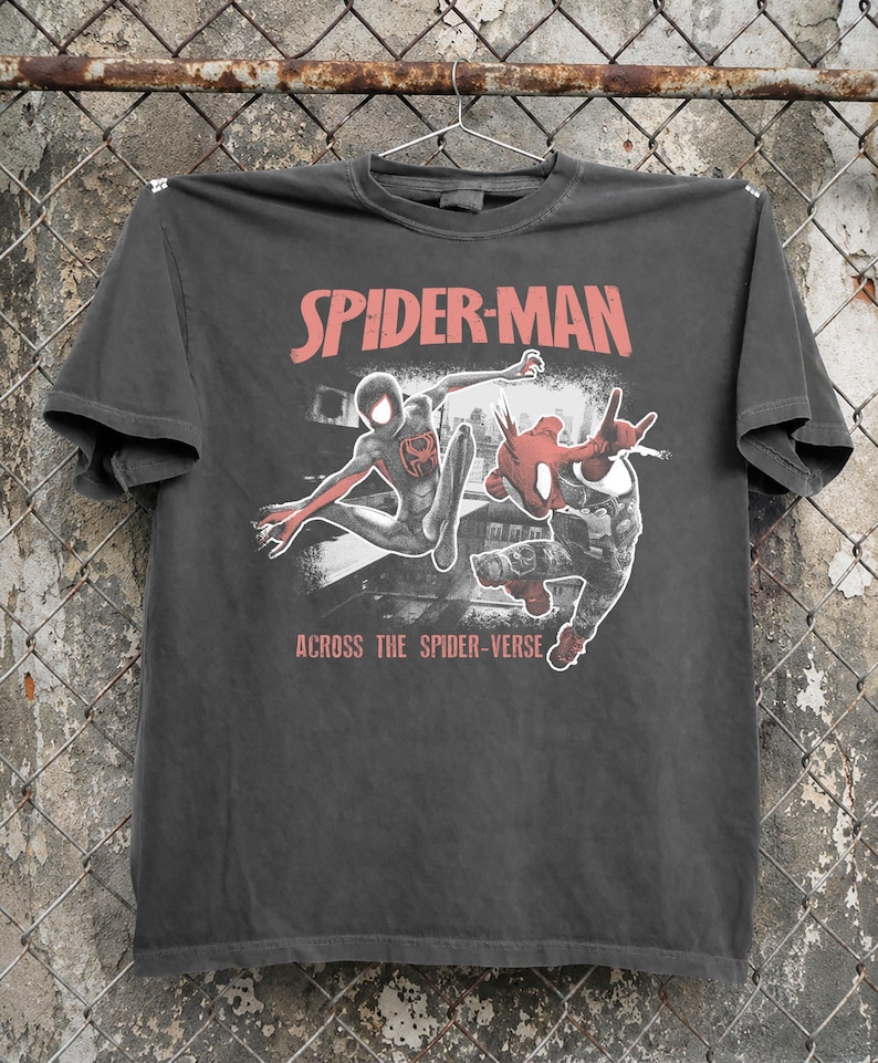Puede incluir: Camiseta gris oscuro con la inscripci&oacute;n roja "SPIDER-MAN" y un gr&aacute;fico de dos personajes de Spider-Man. La frase "ACROSS THE SPIDER-VERSE" est&aacute; impresa debajo.