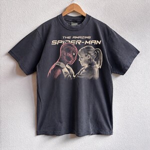 K&ouml;nnte beinhalten: Dunkelgraues T-Shirt mit einer Grafik von "The Amazing Spider-Man". Das Design zeigt Spider-Man und eine Frau, die sich gegen&uuml;berstehen. Der Text "THE AMAZING SPIDER-MAN" ist in Wei&szlig; &uuml;ber der Grafik aufgedruckt. Das T-Shirt h&auml;ngt an einem Holzb&uuml;gel.