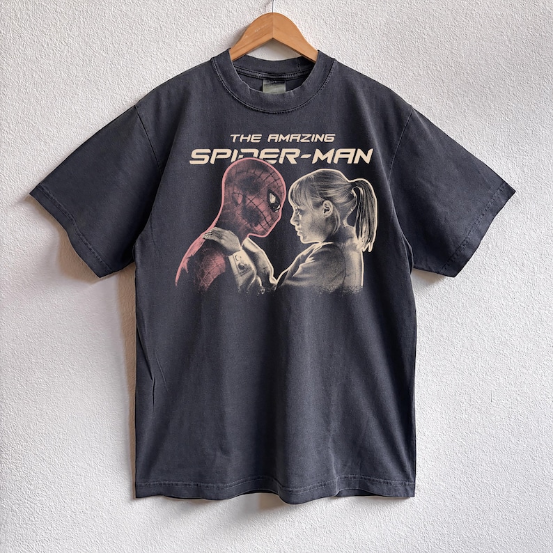 K&ouml;nnte beinhalten: Dunkelgraues T-Shirt mit einer Grafik von "The Amazing Spider-Man". Das Design zeigt Spider-Man und eine Frau, die sich gegen&uuml;berstehen. Der Text "THE AMAZING SPIDER-MAN" ist in Wei&szlig; &uuml;ber der Grafik aufgedruckt. Das T-Shirt h&auml;ngt an einem Holzb&uuml;gel.