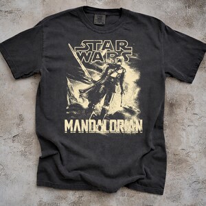 May include: Dark gray t-shirt featuring a distressed graphic of The Mandalorian from Star Wars. The design includes the Star Wars logo and the word "Mandalorian" in a bold font. The shirt has a crew neck and short sleeves.