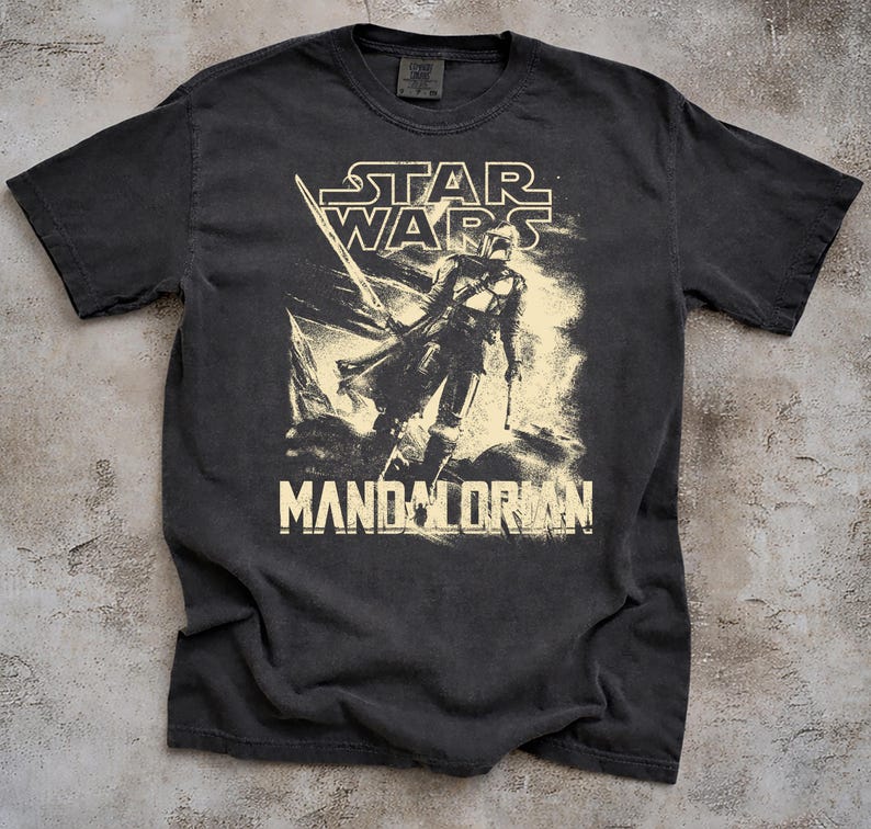 May include: Dark gray t-shirt featuring a distressed graphic of The Mandalorian from Star Wars. The design includes the Star Wars logo and the word "Mandalorian" in a bold font. The shirt has a crew neck and short sleeves.