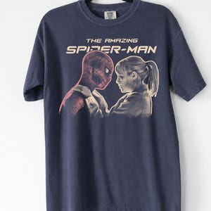 K&ouml;nnte beinhalten: Marineblaues T-Shirt mit einer Grafik von "The Amazing Spider-Man". Das Design zeigt Spider-Man und eine Frau, die sich gegen&uuml;berstehen. Der Text "THE AMAZING SPIDER-MAN" ist im Vintage-Stil &uuml;ber dem Bild gedruckt.