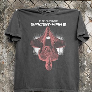 May include: Dark gray t-shirt featuring the text "THE AMAZING SPIDER-MAN 2" in white. The shirt displays an upside-down image of Spider-Man in a red and black suit against a cityscape backdrop.