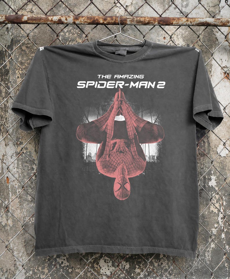 May include: Dark gray t-shirt featuring the text "THE AMAZING SPIDER-MAN 2" in white. The shirt displays an upside-down image of Spider-Man in a red and black suit against a cityscape backdrop.