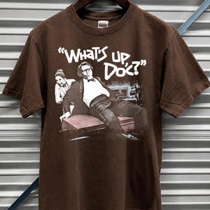 May include: Dark chocolate brown t-shirt featuring a graphic from the movie "What's Up, Doc?" in white text. The image shows a man in a suit and a woman. The shirt is hanging on a wooden hanger.