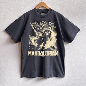 May include: Dark gray t-shirt featuring a graphic of the Mandalorian from Star Wars. The design includes the Star Wars logo and the word "Mandalorian" in a bold font. The shirt is hanging on a wooden hanger.