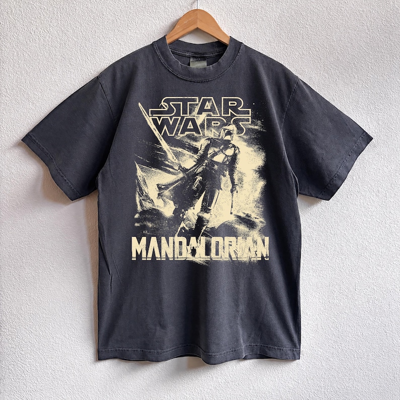 May include: Dark gray t-shirt featuring a graphic of the Mandalorian from Star Wars. The design includes the Star Wars logo and the word "Mandalorian" in a bold font. The shirt is hanging on a wooden hanger.