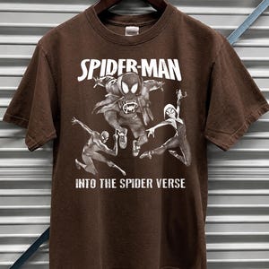 May include: Dark chocolate brown t-shirt featuring a graphic of Spider-Man and other Spider-Verse characters. The text "SPIDER-MAN" and "INTO THE SPIDER VERSE" are printed in white. The shirt is hanging on a wooden hanger.