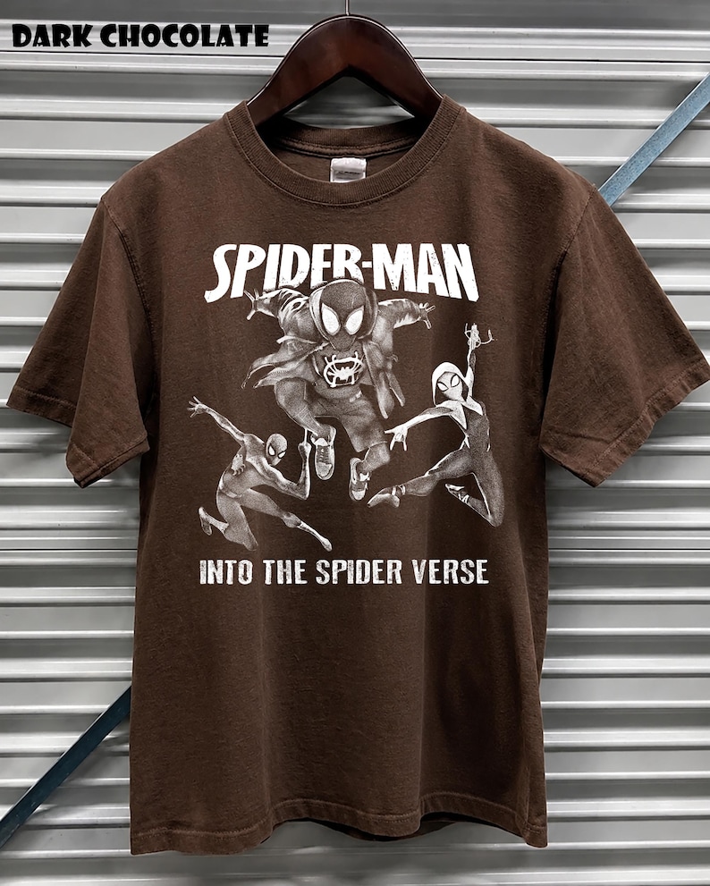 May include: Dark chocolate brown t-shirt featuring a graphic of Spider-Man and other Spider-Verse characters. The text "SPIDER-MAN" and "INTO THE SPIDER VERSE" are printed in white. The shirt is hanging on a wooden hanger.