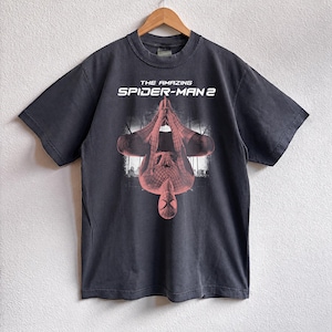 May include: Dark gray t-shirt featuring the text "THE AMAZING SPIDER-MAN 2" above a red and black graphic of Spider-Man hanging upside down. The shirt is hanging on a wooden hanger.