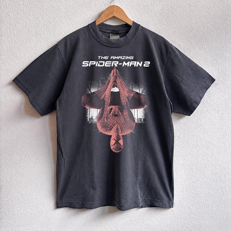 May include: Dark gray t-shirt featuring the text "THE AMAZING SPIDER-MAN 2" above a red and black graphic of Spider-Man hanging upside down. The shirt is hanging on a wooden hanger.