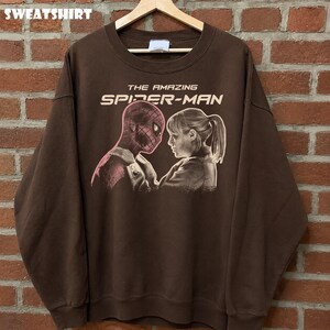 K&ouml;nnte beinhalten: Braunes Sweatshirt mit einem Aufdruck von Spider-Man und einer Frau, mit dem Text "THE AMAZING SPIDER-MAN" in Wei&szlig;. Das Wort "SWEATSHIRT" ist oben aufgedruckt. Das Sweatshirt h&auml;ngt an einem Holzkleiderb&uuml;gel.
