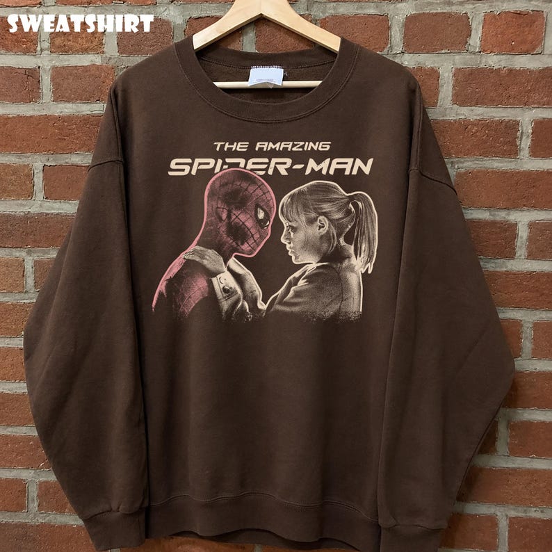 K&ouml;nnte beinhalten: Braunes Sweatshirt mit einem Aufdruck von Spider-Man und einer Frau, mit dem Text "THE AMAZING SPIDER-MAN" in Wei&szlig;. Das Wort "SWEATSHIRT" ist oben aufgedruckt. Das Sweatshirt h&auml;ngt an einem Holzkleiderb&uuml;gel.