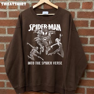 May include: A brown sweatshirt featuring a Spider-Man graphic. The design includes the words "SPIDER-MAN" and "INTO THE SPIDER VERSE" in white, along with illustrations of Spider-Man characters in action poses. The word "SWEATSHIRT" is at the top.