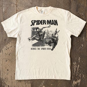 Puede incluir: Camiseta color crema con un dise&ntilde;o en blanco y negro de Spider-Man en acci&oacute;n. El texto "SPIDER-MAN" est&aacute; sobre la imagen y "ACROSS THE SPIDER-VERSE" debajo. Cuello redondo y mangas cortas.